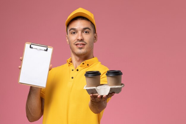 Widok z przodu męski kurier w żółtym mundurze, trzymając notatnik i filiżanki kawy dostawy na różowym biurku