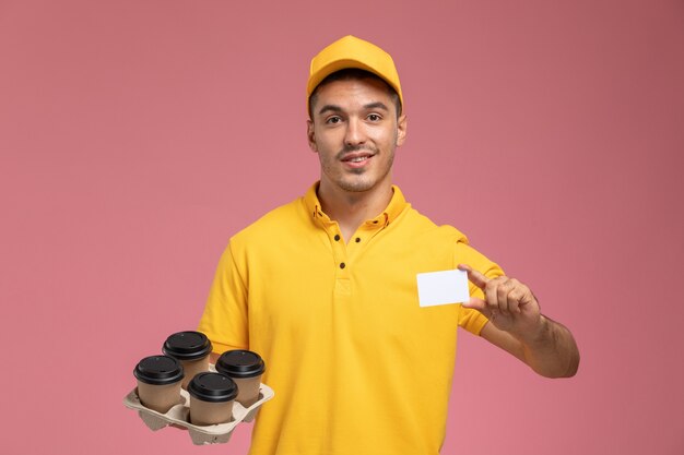 Widok z przodu męski kurier w żółtym mundurze trzymający plastikową kartę i dostawy filiżanek kawy na różowym tle