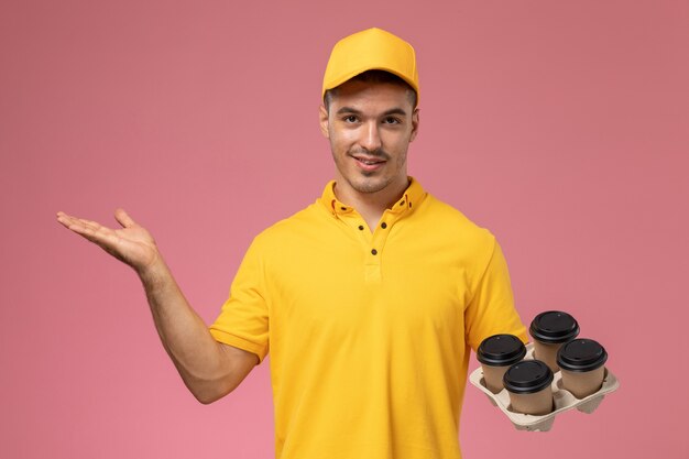 Widok z przodu męski kurier w żółtym mundurze, uśmiechając się i trzymając filiżanki kawy dostawy na różowym biurku