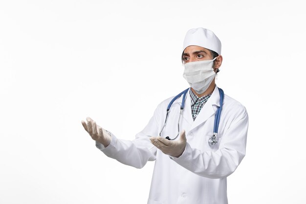 Widok z przodu męski lekarz w białym garniturze i masce z powodu koronawirusa na jasnobiałej powierzchni