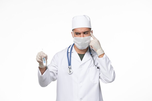 Widok z przodu męski lekarz w kombinezonie medycznym i masce przeciw koronawirusowi trzymającemu urządzenie na białym biurku wirus wirusowy - choroba pandemia