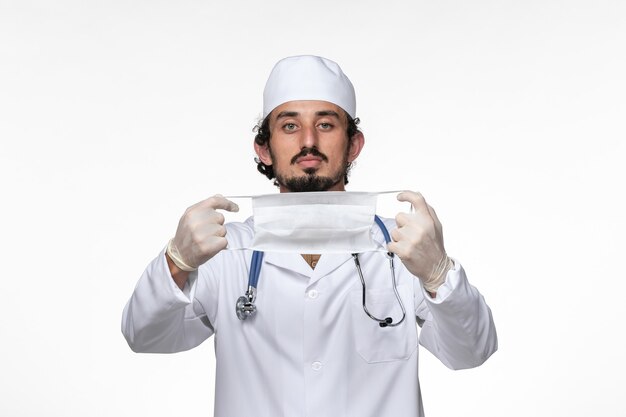 Widok z przodu męski lekarz w kombinezonie medycznym i trzymający maskę jako ochronę przed zakaźną chorobą wirusową na białej ścianie zdrowie pandemia