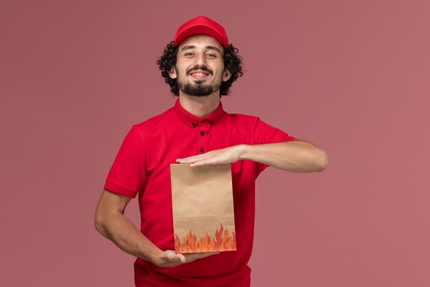 Widok z przodu mężczyzna dostawy kurierskiej mężczyzna w czerwonej koszuli i pelerynie, trzymając papierowy pakiet żywności na różowej ścianie
