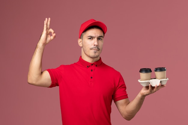 Widok z przodu mężczyzna kurier w czerwonym mundurze, trzymając brązowe filiżanki kawy dostawy na jasnoróżowej ścianie pracownik płci męskiej