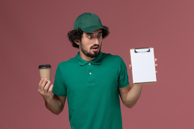 Widok z przodu mężczyzna kurier w zielonym mundurze i pelerynie trzymający dostawę filiżankę kawy z notatnikiem na różowym tle jednolita praca firmy kurierskiej