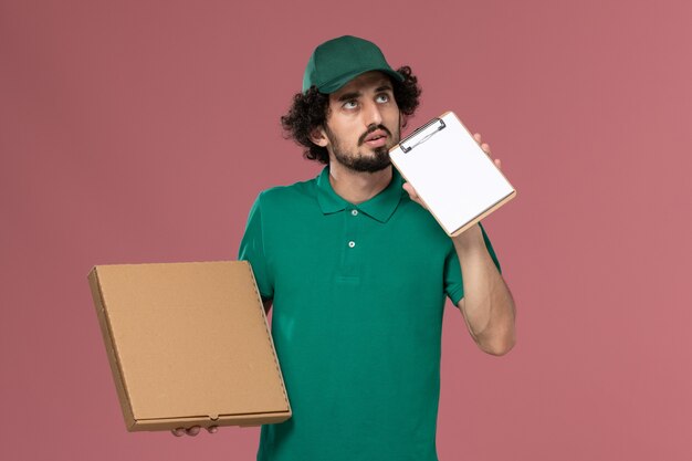 Widok z przodu mężczyzna kurier w zielonym mundurze i pelerynie trzymający dostawę pudełko z jedzeniem i myślący na różowym tle usługa jednolita dostawa praca