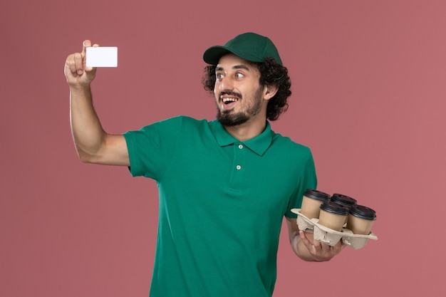 Widok z przodu mężczyzna kurier w zielonym mundurze i pelerynie trzymający filiżanki z kawą z kartą na różowym tle jednolita usługa dostawy