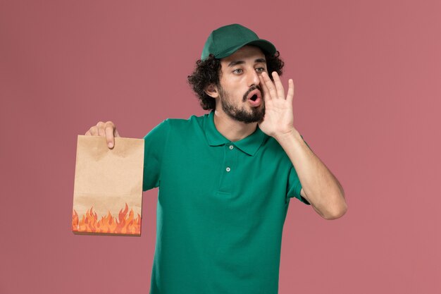 Widok z przodu mężczyzna kurier w zielonym mundurze i pelerynie trzymający papierowy pakiet żywności wzywając na różowym tle pracownik służbowy jednolity praca dostawy