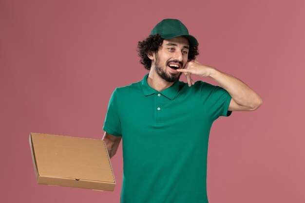 Widok z przodu mężczyzna kurier w zielonym mundurze i pelerynie trzymający pudełko z jedzeniem pokazujący pozę telefoniczną na różowym tle pracownik służbowy mundur dostawy
