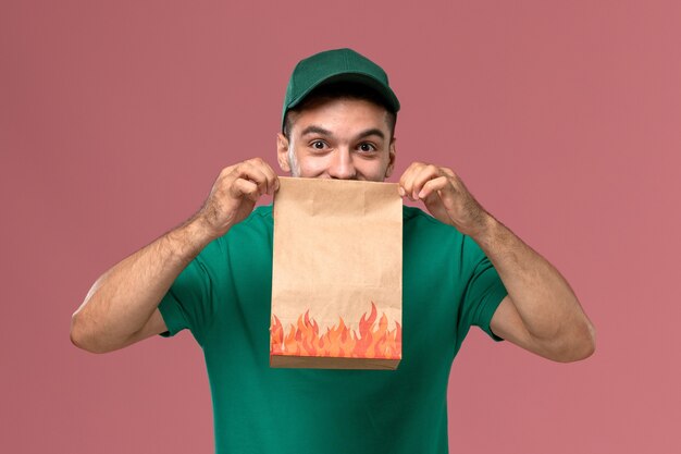 Widok z przodu mężczyzna kurier w zielonym mundurze, trzymając papierowy pakiet żywności i uśmiechając się na różowym tle