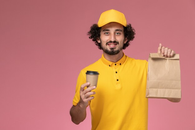 Widok z przodu mężczyzna kurier w żółtym mundurze i pelerynie trzymający dostawę kubek kawy opakowanie żywnościowe na różowej ścianie