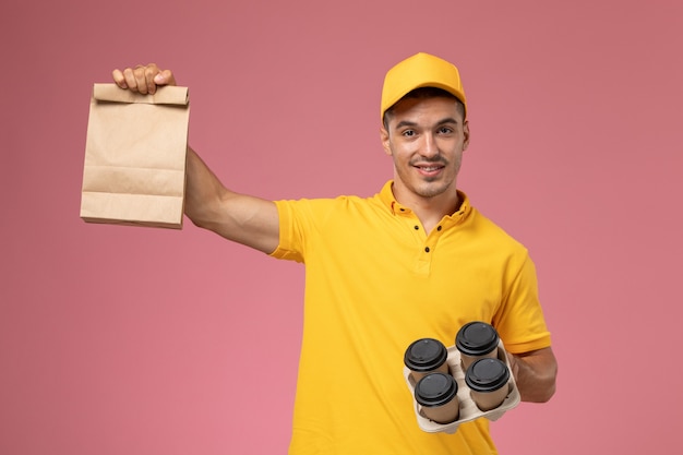 Widok z przodu mężczyzna kurier w żółtym mundurze, trzymając pakiet żywności i filiżanki kawy dostawy, uśmiechając się na różowym tle