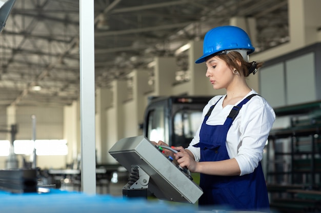 Widok z przodu młoda atrakcyjna dama w niebieskim kolorze skafandra i hełmie kontrolującym maszyny w hangarze pracującym podczas budowy architektury budynków w ciągu dnia
