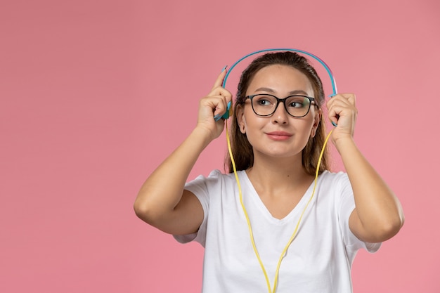 Widok z przodu młoda atrakcyjna kobieta w białej koszulce pozowanie do słuchania muzyki w słuchawkach smi na różowym tle