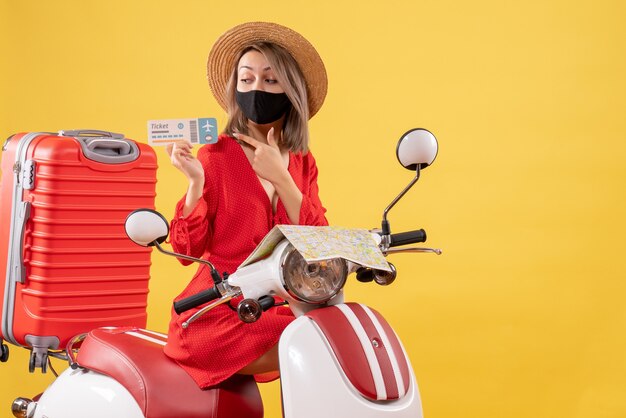 Widok z przodu młoda dama z czarną maską na motorowerze z czerwoną walizką wskazującą na bilet