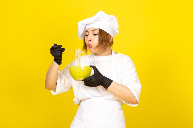 Widok z przodu młoda kobieta kucharz w białym garniturze i białej czapce w czarnych rękawiczkach, trzymając zielony talerz, mieszając spaghetti na żółtym