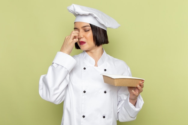 Widok Z Przodu Młoda Kobieta Kucharz W Białym Garniturze I Czapce Trzymającej Pakiet Zamykający Nos Na Zielonej ścianie Pani Pracuje Kolor Kuchni żywności