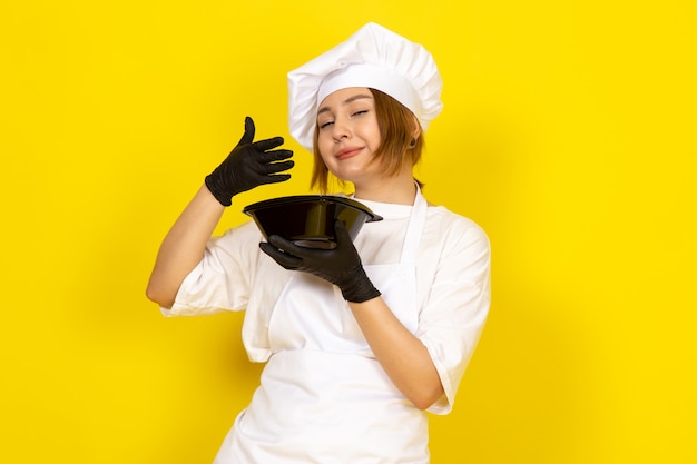 Widok z przodu młoda kobieta kucharz w białym garniturze kucharza i białej czapce w czarnych rękawiczkach trzyma czarną miskę uśmiechając się na żółtym