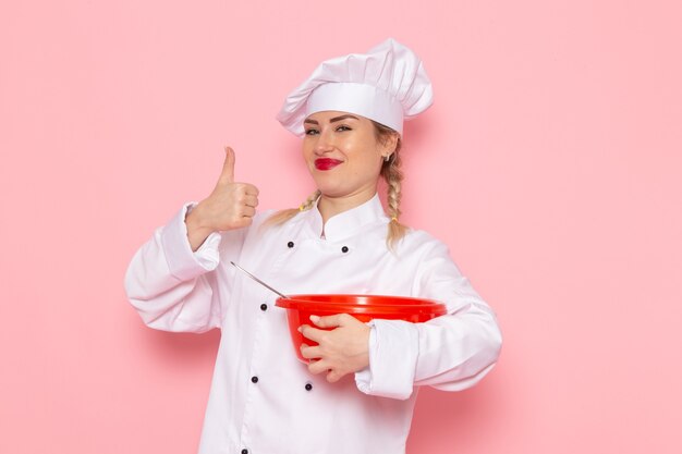 Widok Z Przodu Młoda Kobieta Kucharz W Białym Garniturze Trzymając Czerwoną Miskę Uśmiechając Się Na Różowym Miejscu Gotować Kuchni Praca Praca Zdjęcie