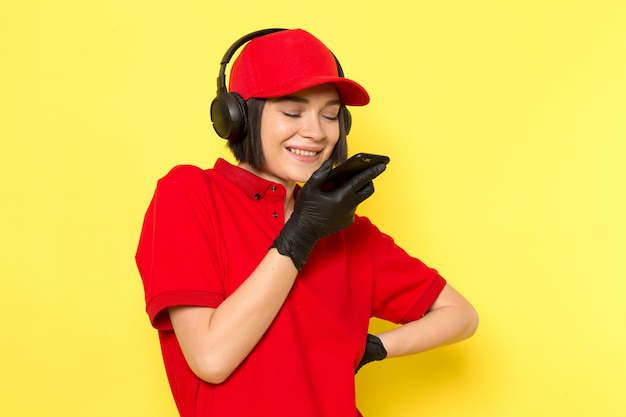 Widok z przodu młoda kobieta kurier w czerwone jednolite czarne rękawiczki i czerwoną czapkę w słuchawkach wysyłających wiadomość głosową