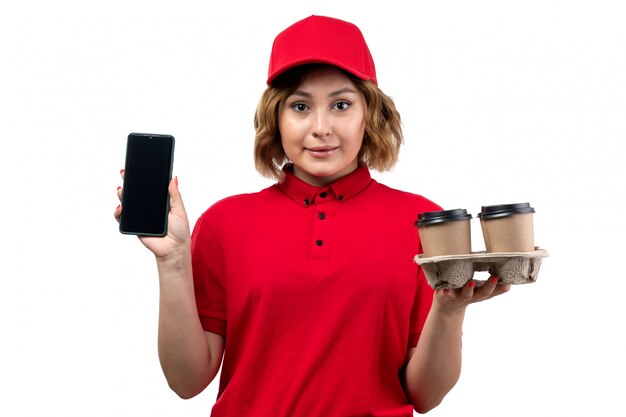Widok z przodu młoda kobieta kurier w czerwonej koszuli czerwona czapka, trzymając smartfon i filiżanki do kawy