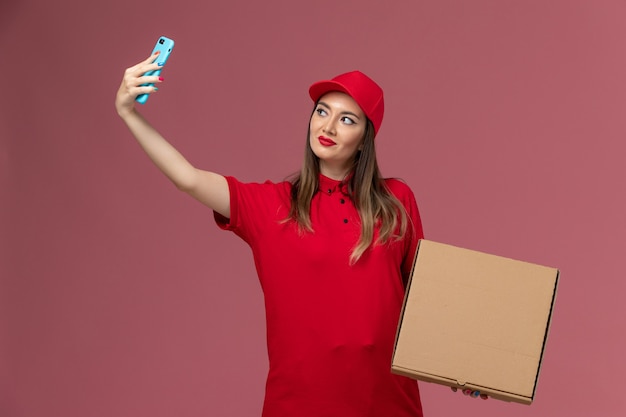 Widok z przodu młoda kobieta kurier w czerwonym mundurze, trzymając pudełko z dostawą żywności i robiąc zdjęcie z nim na różowym tle firma dostarczająca mundur