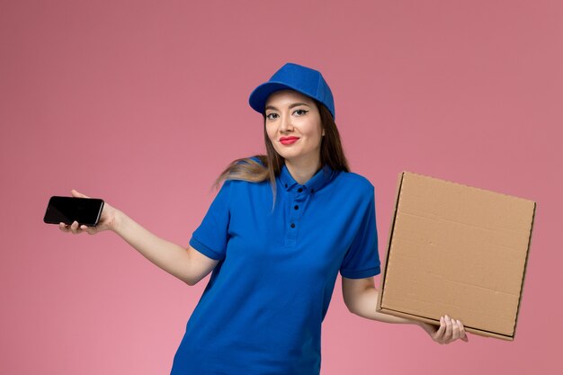 Widok Z Przodu Młoda Kobieta Kurier W Niebieskim Mundurze I Pelerynie, Trzymając Pudełko Z Jedzeniem I Jej Telefon Na Różowej ścianie