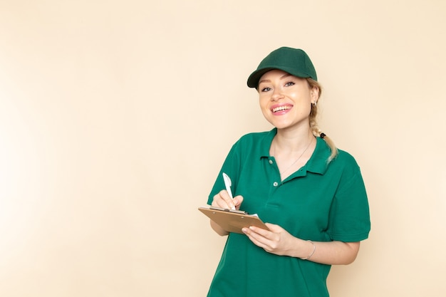 Widok z przodu młoda kobieta kurier w zielonym mundurze i zielonej pelerynie zapisując notatki uśmiechając się na lekkiej przestrzeni pracy kobiety mundurze
