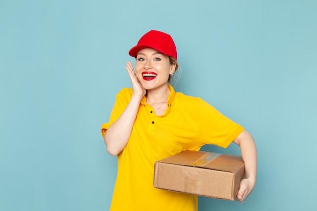 Widok z przodu młoda kobieta kurier w żółtej koszuli i czerwonej pelerynie uśmiechając się i trzymając pakiet na niebieskim pracowniku pracy przestrzeni