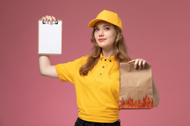 Widok z przodu młoda kobieta kurier w żółtym mundurze, trzymając pakiet żywności dostawy i mały notatnik na różowym biurku jednolity pracownik usługi dostawy
