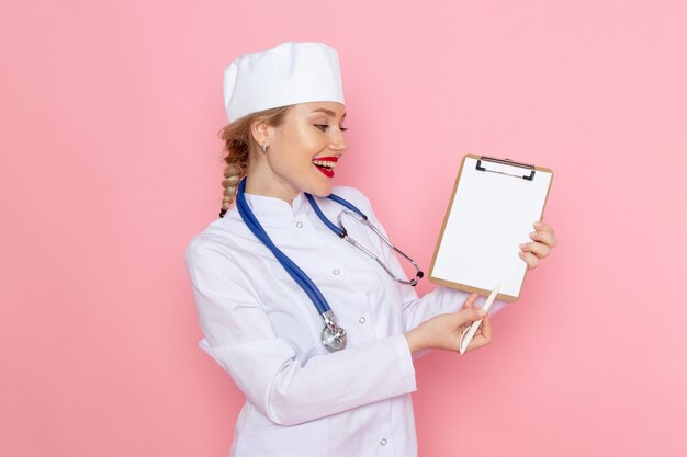 Widok z przodu młoda kobieta lekarz w białym garniturze medycznym z niebieskim stetoskopem trzymając notatnik z uśmiechem na różowej przestrzeni medycyny pracy pracownika szpitala medycznego