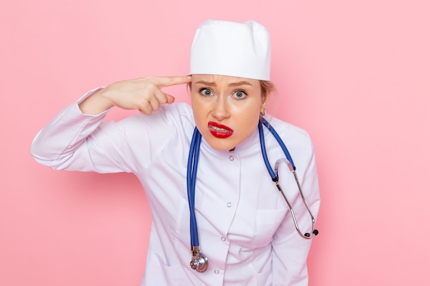 Widok z przodu młoda kobieta lekarz w białym garniturze z niebieskim stetoskopem stwarzających z zabawnym wyrazem na różowej przestrzeni lekarza szpitalnej pracy kobiet
