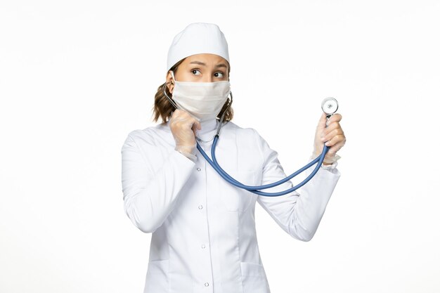 Widok z przodu młoda kobieta lekarz ze sterylną maską z powodu koronawirusa za pomocą stetoskopu na białej powierzchni