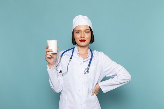 Widok z przodu młoda kobieta pielęgniarka w białym garniturze medycznym i niebieskim stetoskopie trzymając plastikowe szkło na niebieskim biurku lekarz szpitala