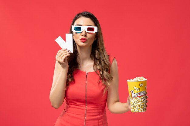 Widok z przodu młoda kobieta trzyma popcorn z biletami d okulary przeciwsłoneczne na jasnoczerwonej powierzchni