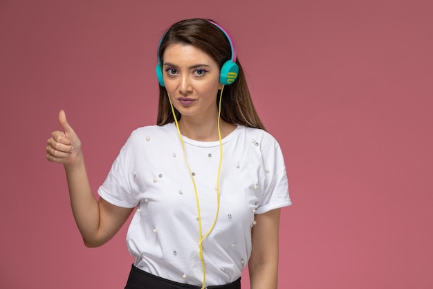 Bezpłatne zdjęcie widok z przodu młoda kobieta w białej koszuli słuchanie muzyki przez słuchawki na różowej ścianie, kolor kobieta stanowi modelka