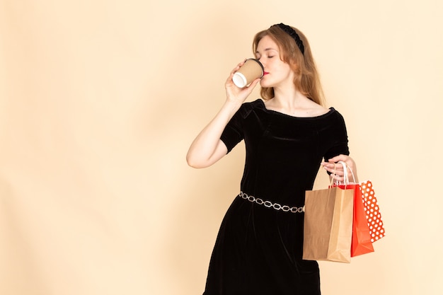 Widok Z Przodu Młoda Kobieta W Czarnej Sukni Z Paskiem łańcuchowym, Trzymając Pakiety Zakupów I Pijąc Kawę Na Beżowym Tle