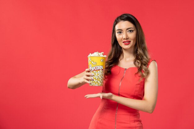 Widok z przodu młoda kobieta w czerwonej koszuli trzymając pakiet popcornu i uśmiechając się na czerwonej powierzchni