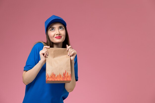 Widok z przodu młoda kurierka w niebieskim mundurze i pelerynie trzymająca papierowe opakowanie żywności na jasnoróżowej ścianie
