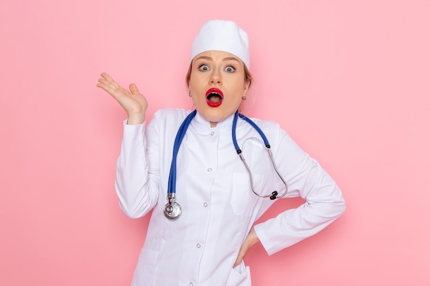 Widok z przodu młoda lekarka w białym garniturze z niebieskim stetoskopem pozuje z zszokowanym wyrazem twarzy na różowej przestrzeni medycyna szpital medyczny lekarz praca kobiet