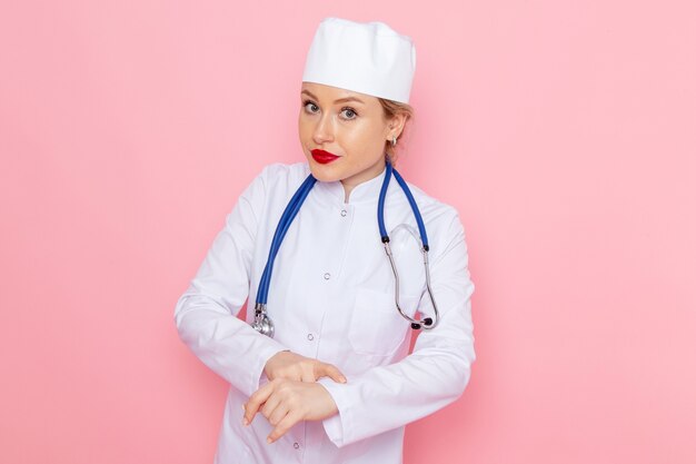 Widok z przodu młoda lekarka w białym garniturze z niebieskim stetoskopem, wskazująca na nadgarstek na różowej przestrzeni medycyna szpital medyczny lekarz praca kobiet