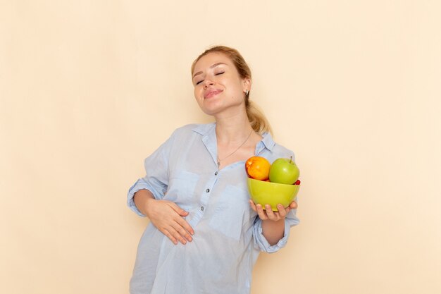 Widok z przodu młoda piękna kobieta w koszuli, trzymając talerz z owocami i uśmiechając się, pozowanie na ścianie kremu owoc model kobiety stanowią dama