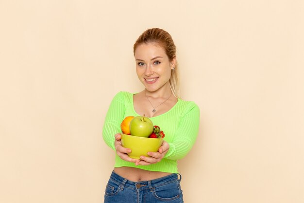Widok z przodu młoda piękna kobieta w zielonej koszuli trzymając talerz z owocami uśmiechając się na lekkiej ścianie kremu owoc model kobieta stanowią dama