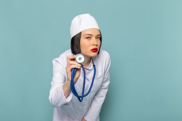 Widok z przodu młoda pielęgniarka w białym garniturze medycznym i niebieskim stetoskopem stwarzających na niebieskim biurku lekarz szpitala