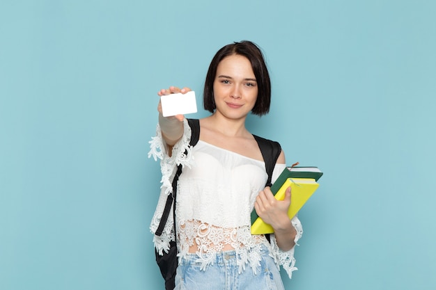 Widok z przodu młoda studentka w białej koszuli, niebieskie dżinsy i czarną torbę, trzymając zeszyty i karty na niebieskiej przestrzeni studentka