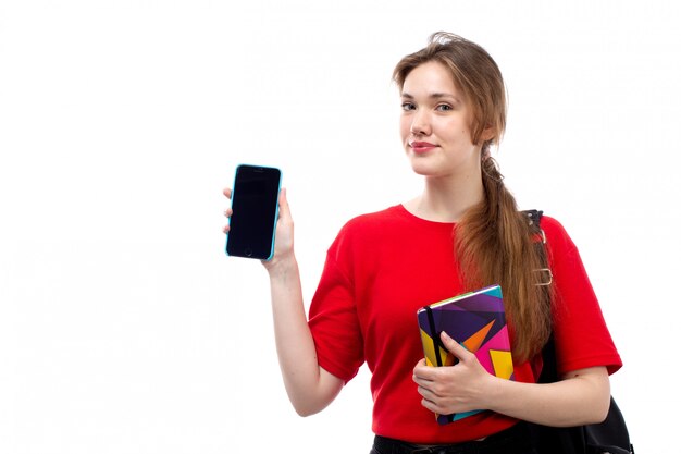 Widok z przodu młoda studentka w czerwonej koszuli czarna torba gospodarstwa zeszyty plików uśmiecha się trzymając jej telefon na białym tle