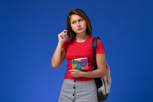 Widok z przodu młoda studentka w czerwonej koszuli na sobie plecak trzymając zeszyt myśli na niebieskim tle.