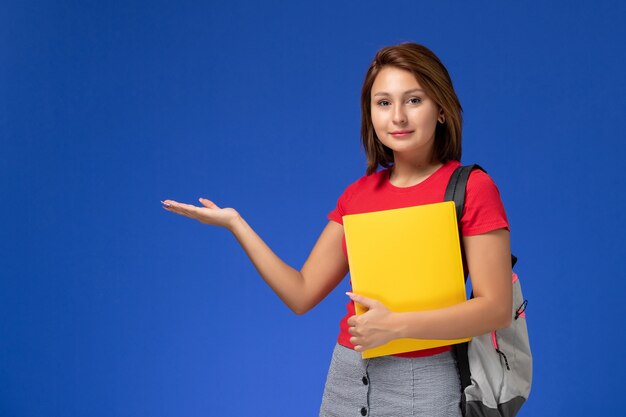 Widok z przodu młoda studentka w czerwonej koszuli z plecakiem, trzymając żółte pliki na jasnoniebieskim tle.