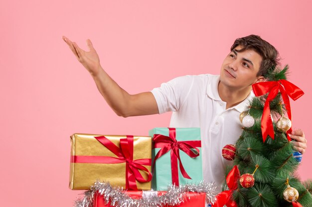 Widok z przodu młodego człowieka wokół świątecznych prezentów i choinki na różowej ścianie