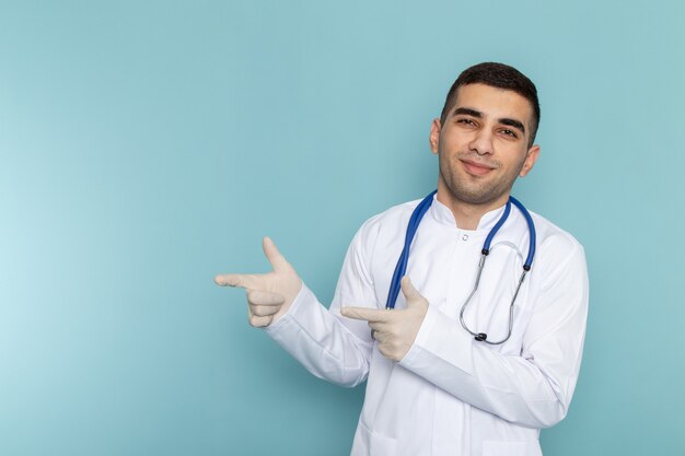 Widok z przodu młodego lekarza płci męskiej w białym garniturze z niebieskim stetoskopem, uśmiechając się i wskazując palcem